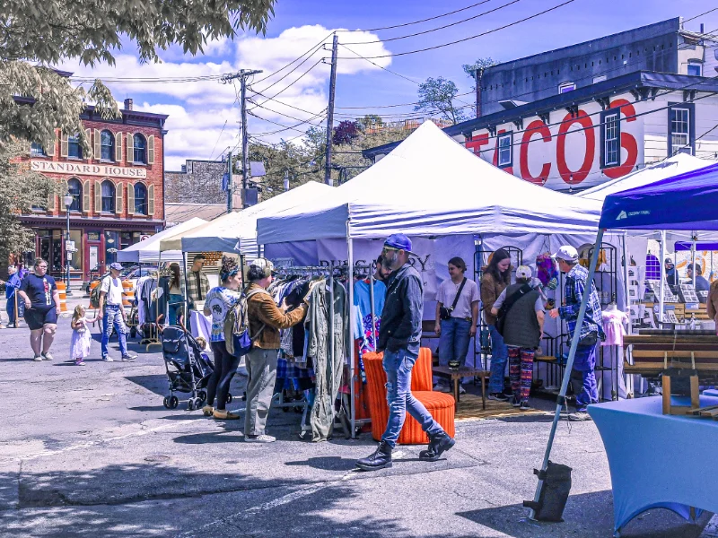 A vibrant outdoor craft market scene with white vendor tents lining a sunny street. Shoppers browse through racks of clothing and handcrafted items, engaging with vendors. A red brick building labeled "Standard House" and a large "TACOS" sign on another building are visible in the background. Families with children and individuals create a lively, bustling atmosphere under a bright blue sky.